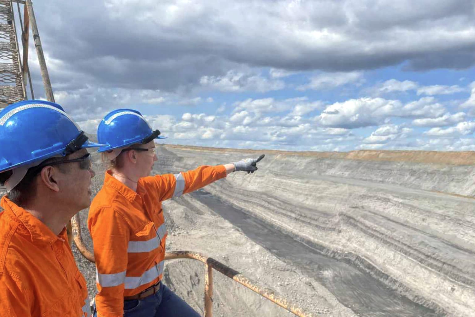 Two mine workers in orange hi-vis and hard hats stand at the edge of an open-cut coal pit, one pointing across the exposed strata of the pit wall.